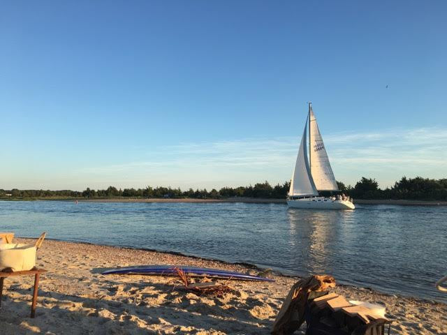 White sailboat on calm blue water at golden hour with sandy beach and driftwood in the foreground, East Hampton