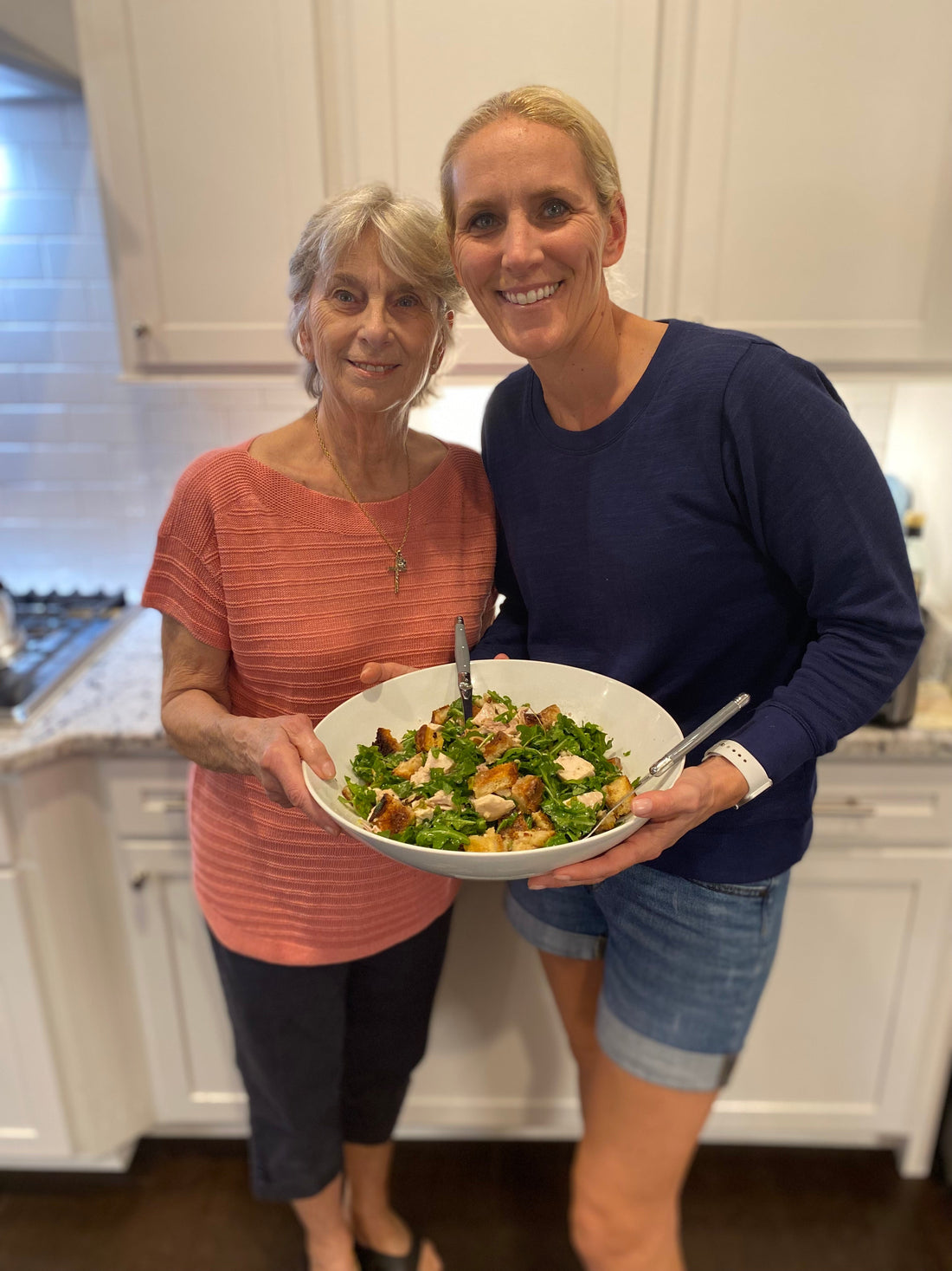 Barbara and Cassandra holding a large bowl of roast chicken with bread and arugula salad in a white kitchen