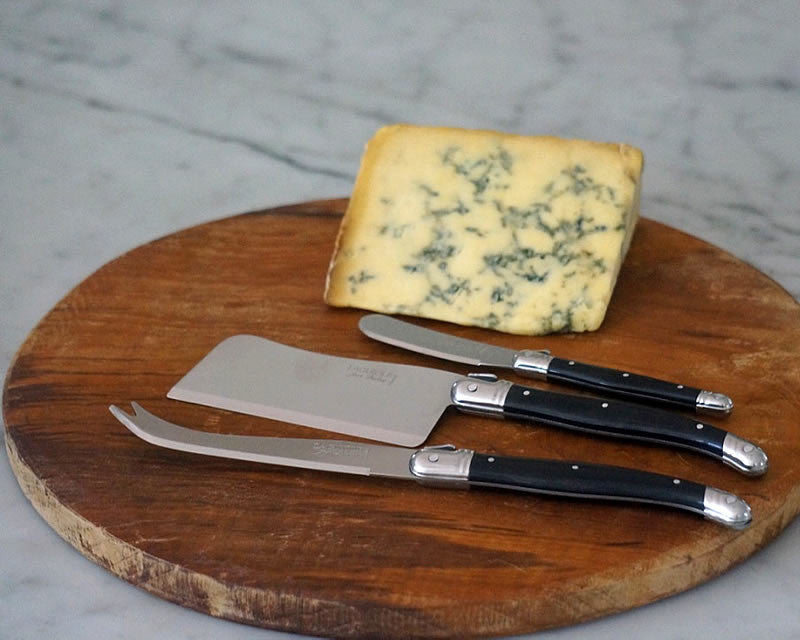 Laguiole black-handle cheese knife set on a round wood board with a wedge of blue cheese on a marble surface