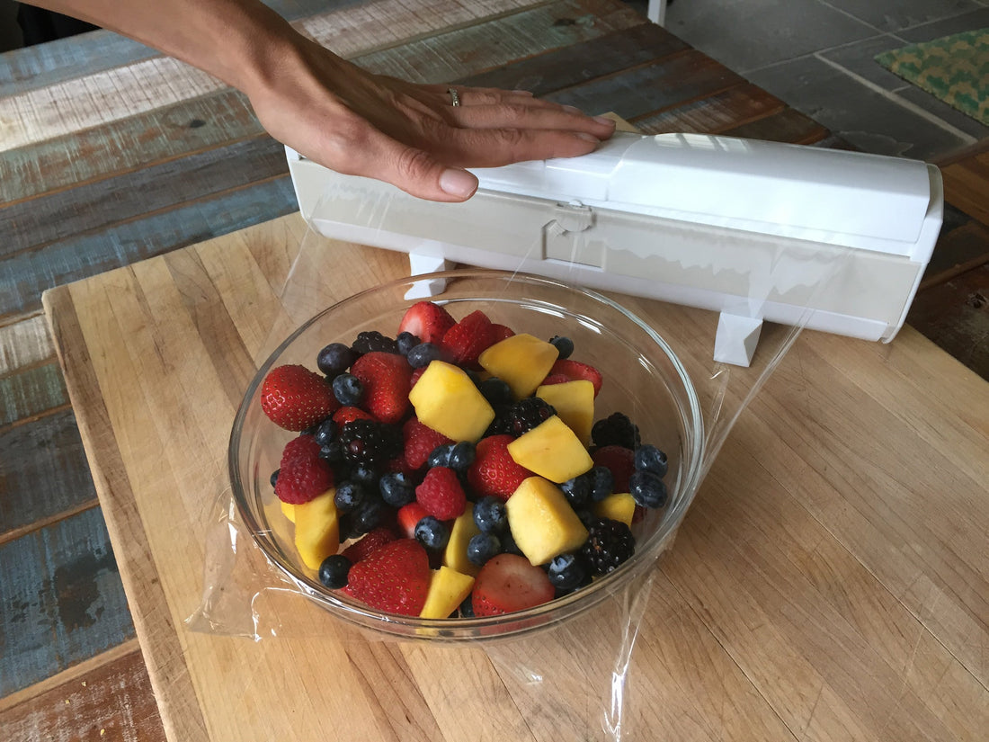 Hand using a plastic wrap dispenser to cover a glass bowl of fresh mixed berries and mango on a cutting board