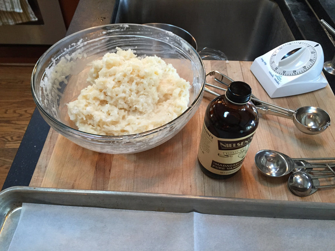 Coconut macaroon batter in a glass mixing bowl with Nielsen-Massey vanilla extract and stainless measuring spoons
