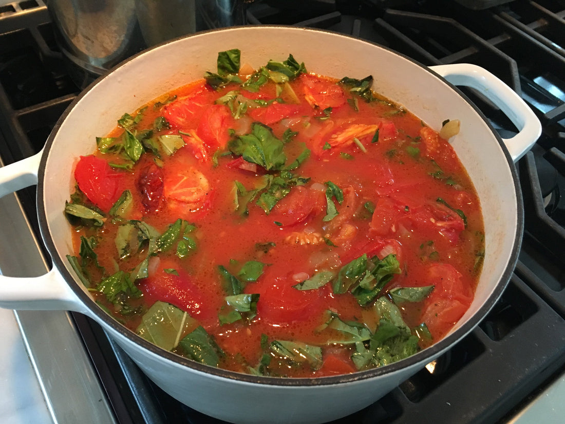 Fresh tomato basil soup simmering with whole tomatoes and basil in a white enamel Dutch oven on a gas stove