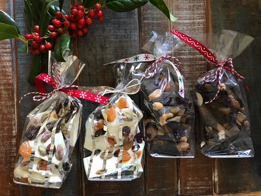 Four holiday gift bags with red and white ribbon containing chocolate bark and mixed nuts on a rustic wood surface