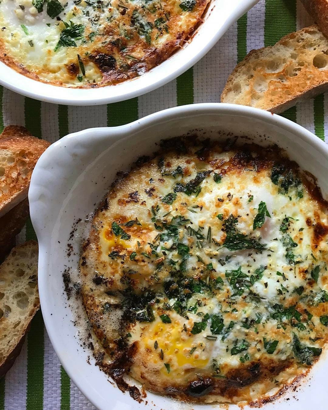 Baked eggs in white ceramic ramekins with fresh herbs and toasted bread on a green striped surface