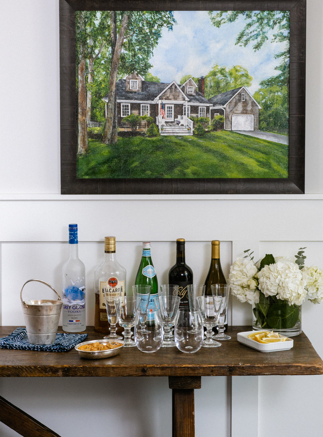 Home bar setup on a rustic wood console with spirit bottles and crystal glasses beneath a house painting