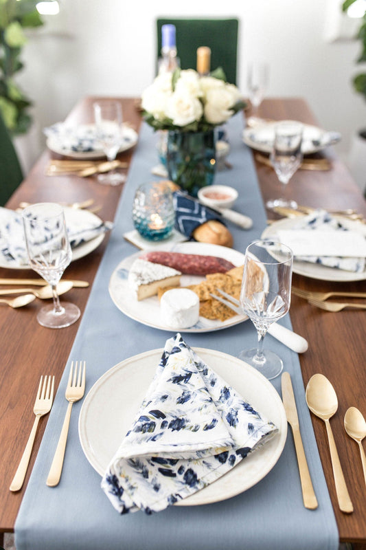 Elegant dinner table setting with blue runner, floral print napkins, gold flatware, crystal stemware, and white rose centerpiece