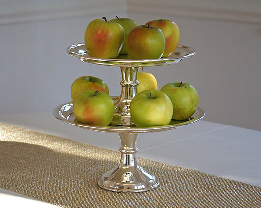 Polished silver two-tiered serving stand filled with green apples on a burlap table runner