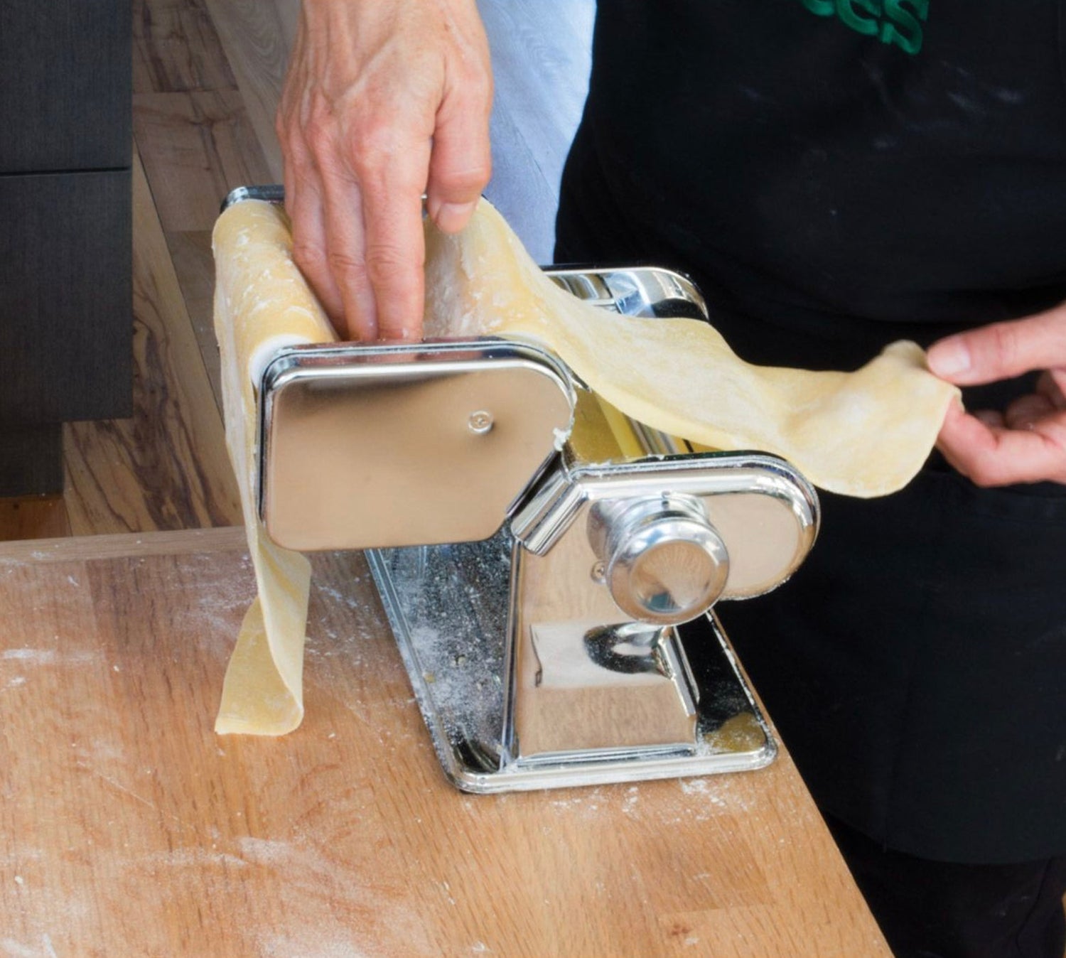 Hands feeding fresh pasta dough through a chrome pasta maker machine on a flour-dusted wooden countertop