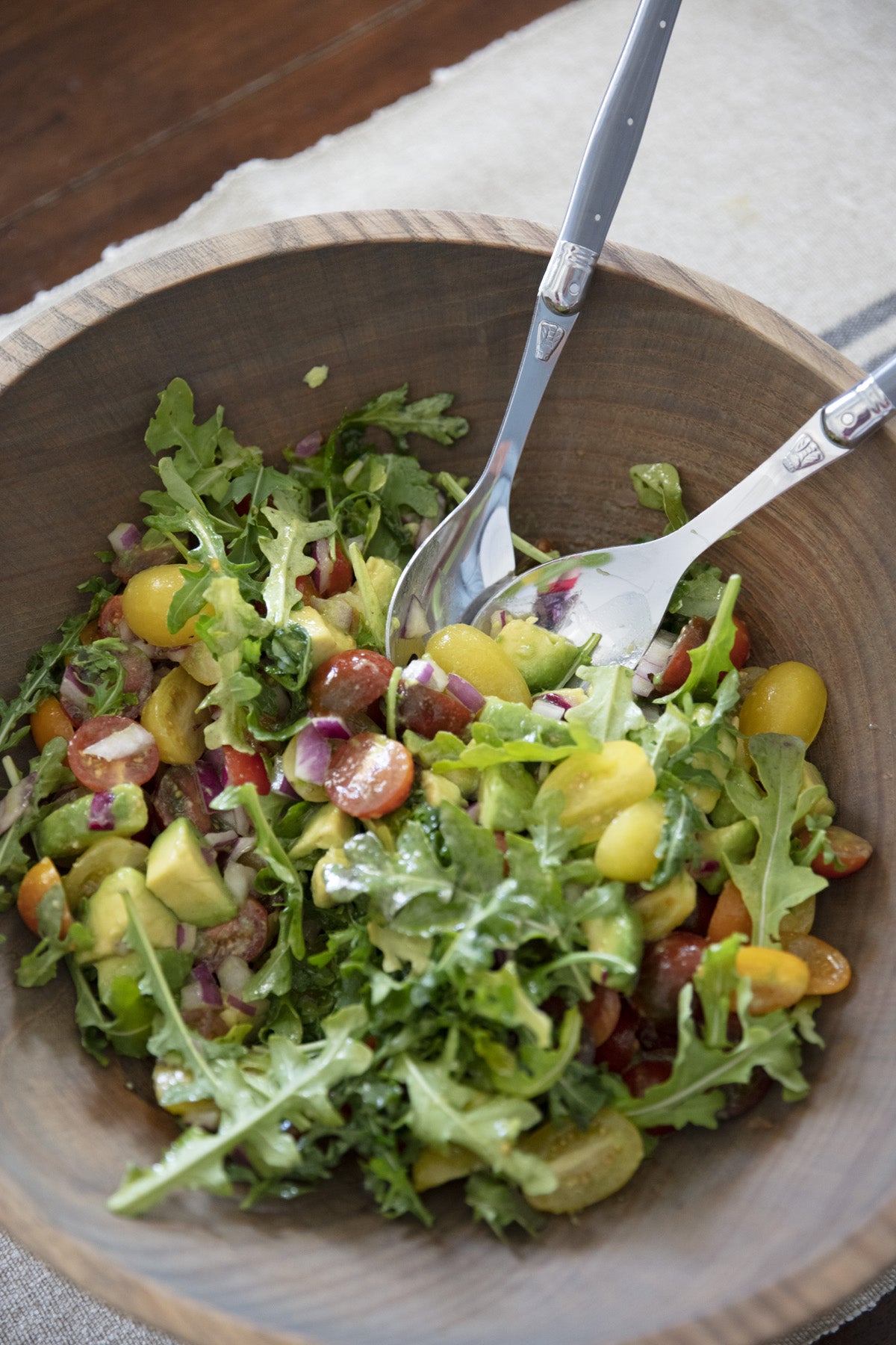 Fresh arugula and tomato salad with avocado in a large wooden bowl with silver serving spoons