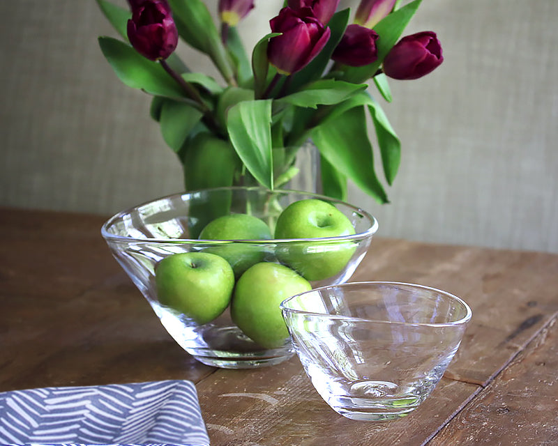 Large clear glass bowl with green apples and a small glass bowl on a rustic wood table, purple tulip bouquet in background