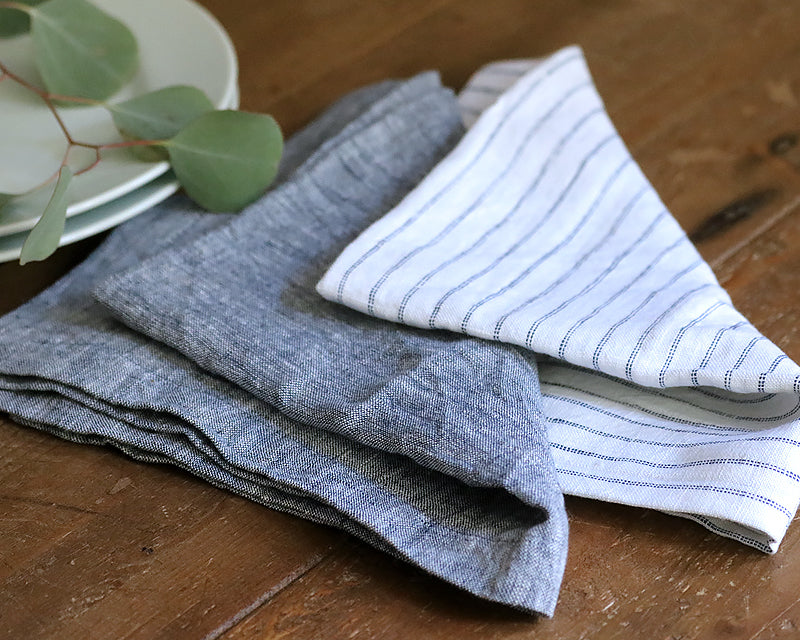 Folded grey linen and white striped cotton napkins on a dark wood table with eucalyptus and white plates