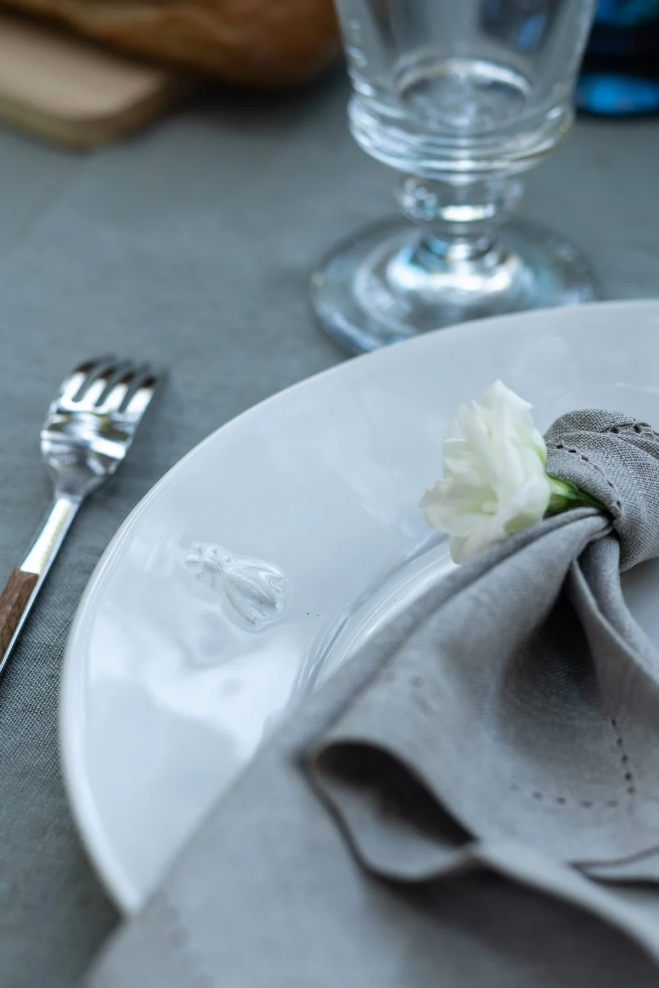 Elegant place setting with white plate featuring bee emblem, grey linen napkin, silver fork, and stemmed wine glass on a grey table