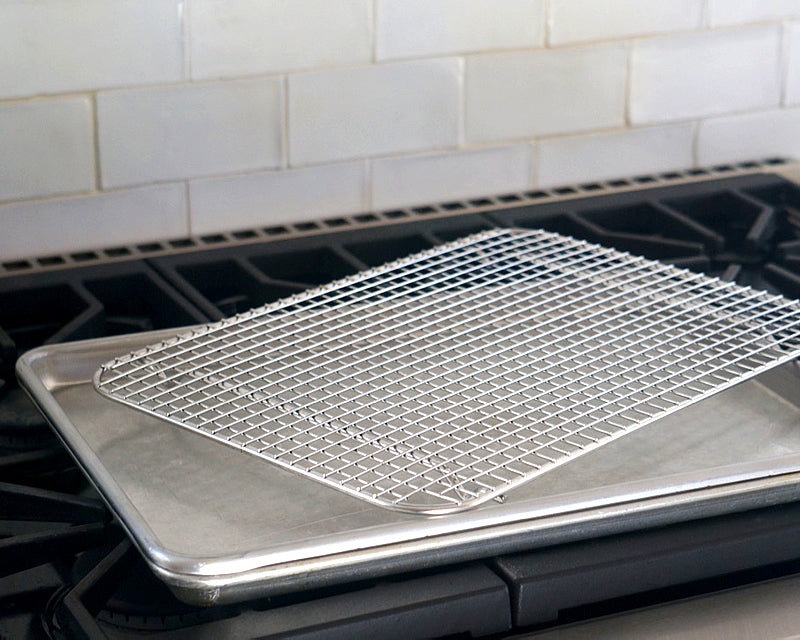 Stainless steel half sheet pan with wire cooling rack on a gas stovetop, white subway tile backsplash