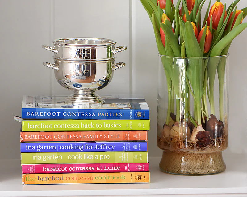 Polished silver footed bowl on top of a stack of Barefoot Contessa cookbooks beside a glass vase with orange tulips