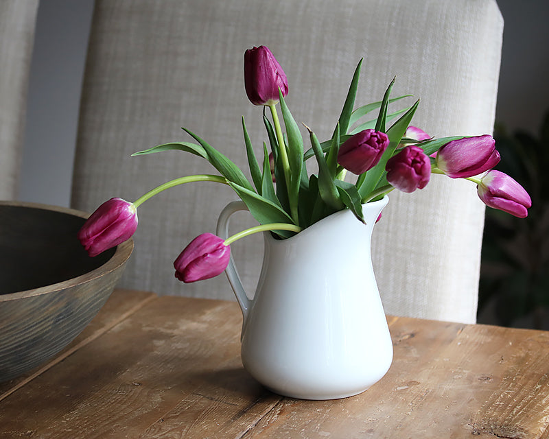 White ceramic milk jug vase with purple tulips on a rustic wood table beside a dark wooden bowl