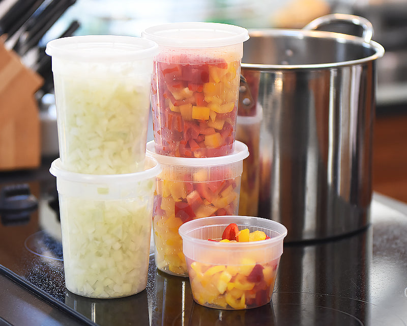 Clear deli-style food storage containers filled with diced onions and colorful peppers beside a stainless steel stockpot on a dark stovetop