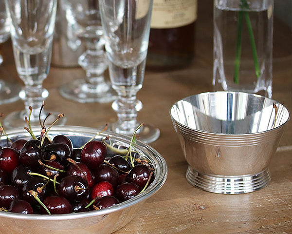 HÔTEL Silver Deco Bar Bowl on a farm table next to the HÔTEL Silver Round Dish filled with cherries.