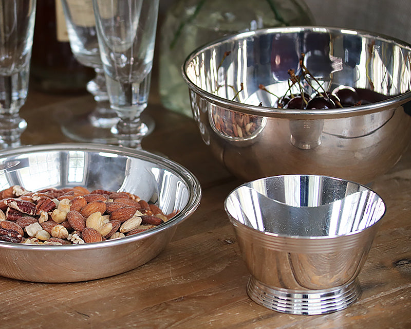 HÔTEL Silver Deco Bar Bowl on a farm table next to the HÔTEL Silver Round Dish filled with assorted nuts.