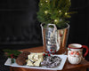 Silver ice bucket holding a Christmas tree decoration displayed next to a platter of holiday treats and a Santa mug.