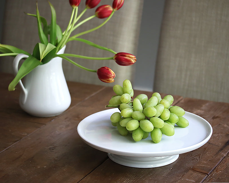 White cake stand by Pillivuyt sits on a wood board with a pile of green grapes. 