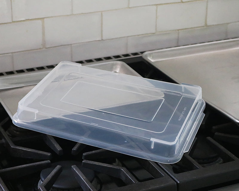 Quarter sheet pan cover sits diagonally on top of the quarter sheet pan and next to a half sheet pan on a stove. 