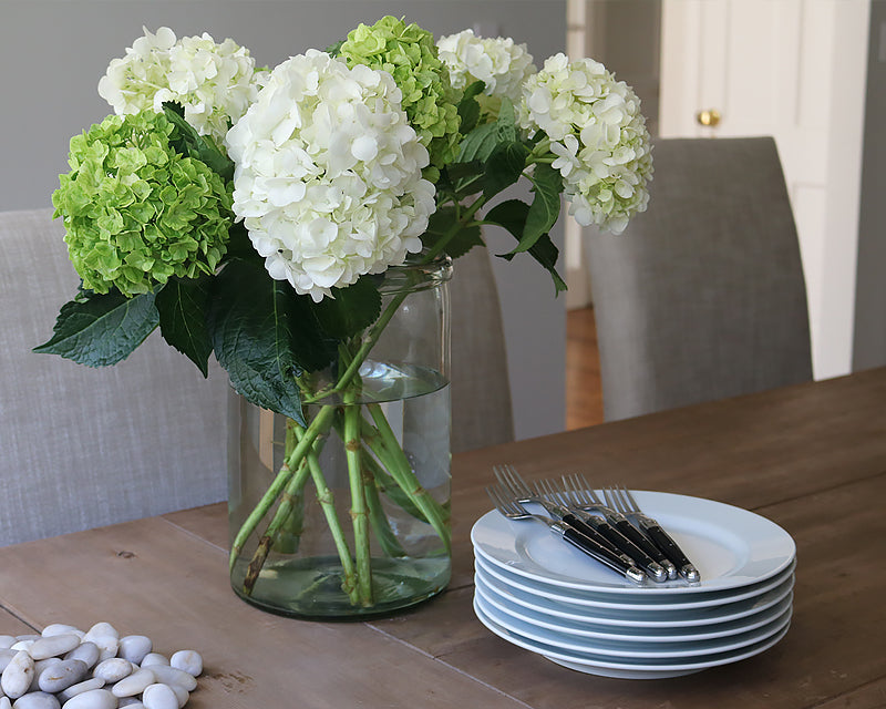 A stack of white Dessert Plates sit on a farm table with our everyday flatware in black. A vase of hydrangeas brightens up this simple table. 