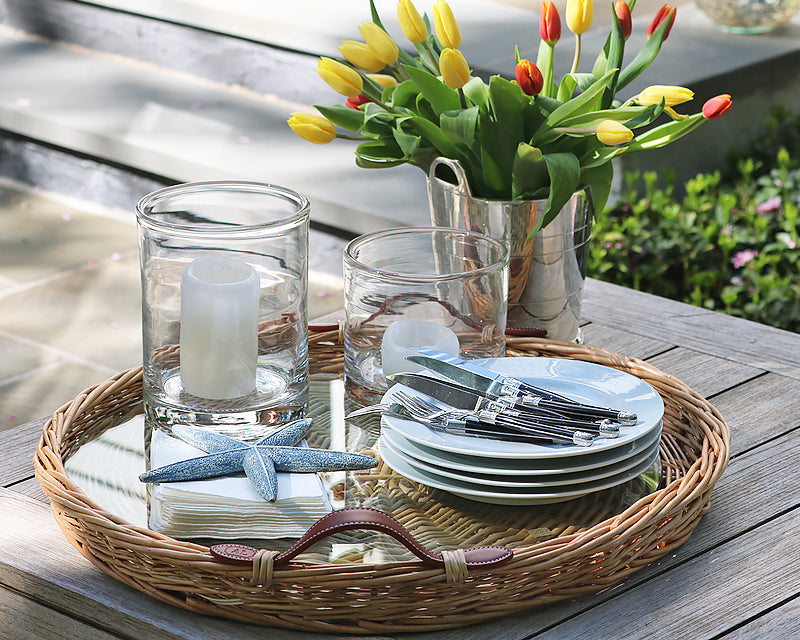 Beautiful outdoor setting ready for a party that includes our Woven Serving Tray, white dinnerware dishes, Everyday flatware, Clear Glass Hurricane Lamps and our Hotel Silver Wine Chiller filled with fresh cut flowers